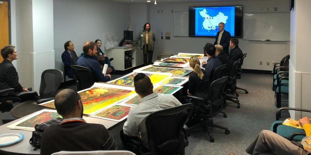 Image of a group of adults sitting around a table in a conference room. The table has colorful artwork on it, and there are two presenters at the front of the room near a projected slide depicting a map of china.
